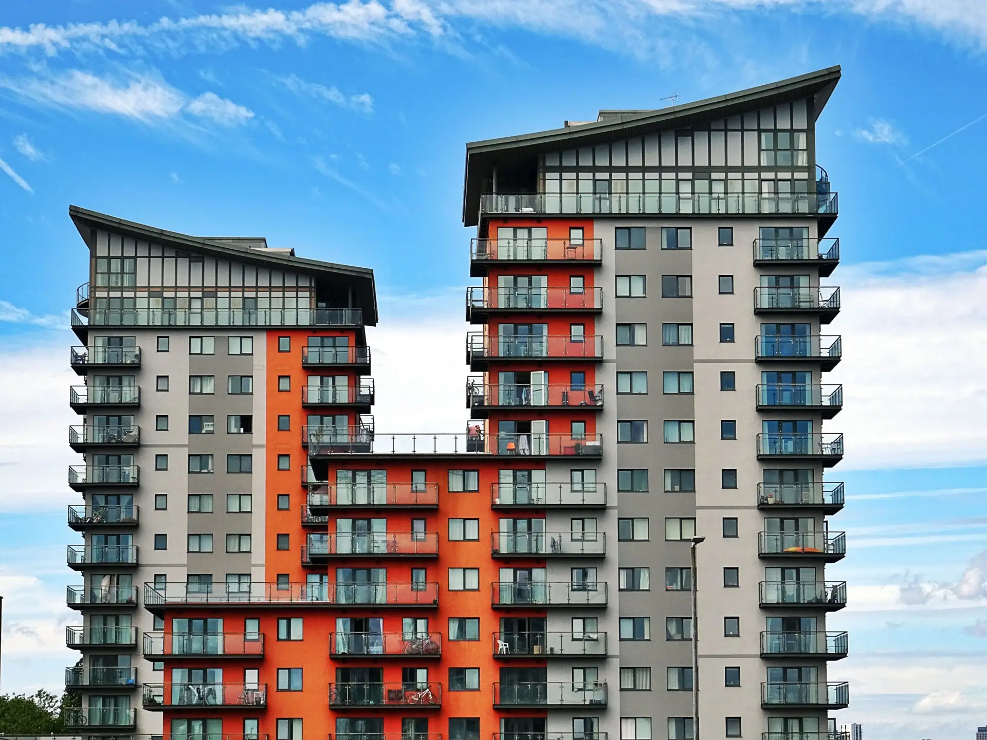 Two modern, multi-story apartment buildings with red and gray facades, many balconies, and large windows offer residents property-wide WiFi, all set against a blue sky with wispy clouds.