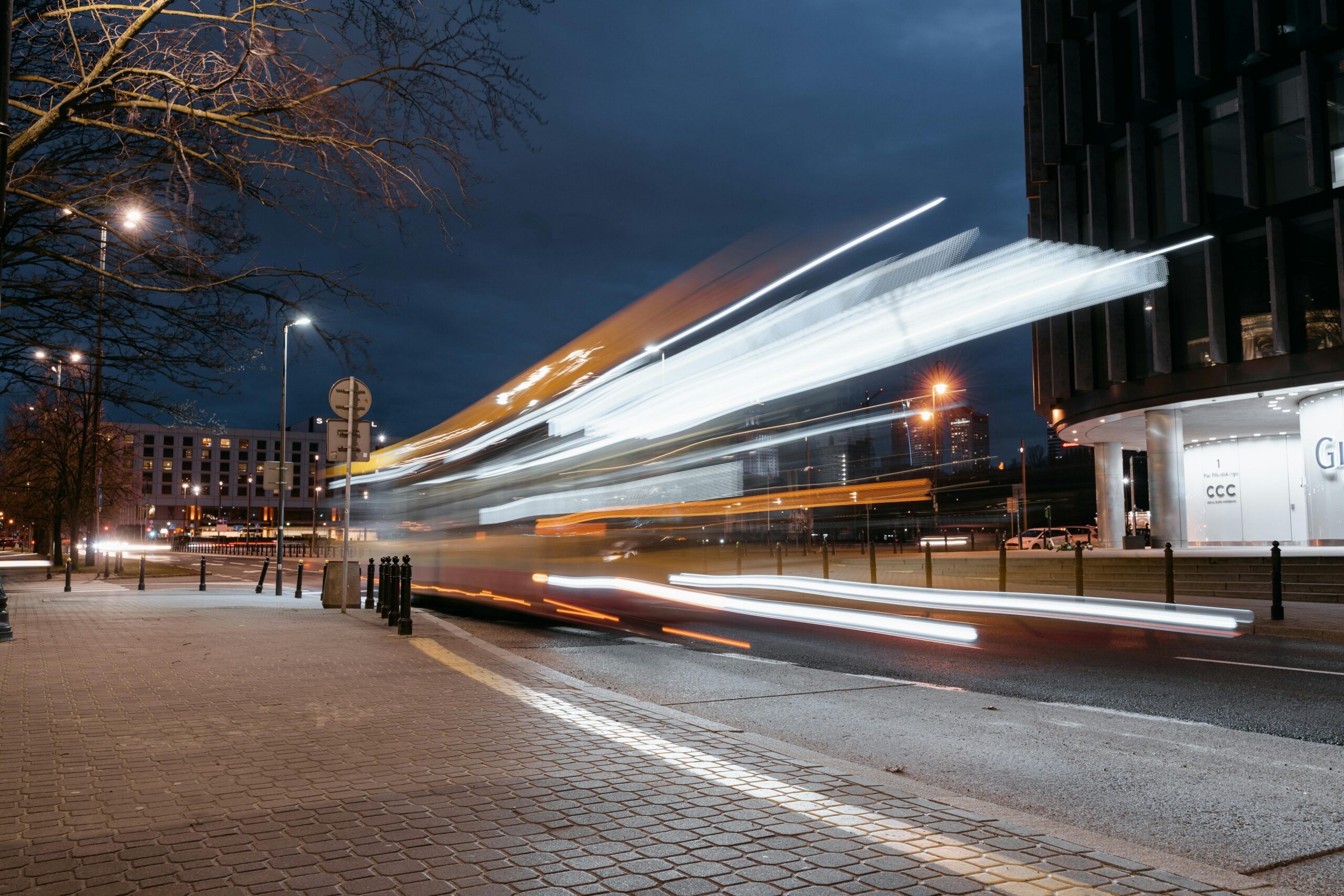 A blurred city bus with light trails speeds down a street at dusk, passing modern buildings and glowing streetlights—like messages racing through a voicemail menu—beneath dark evening clouds.