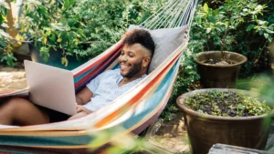 A person smiles while using a laptop in a colorful hammock surrounded by lush green plants and large pots, enjoying a relaxing outdoor setting.