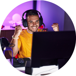 A young man wearing headphones and a yellow shirt smiles and raises his fists in excitement while sitting at a computer desk, likely celebrating a win while gaming. The room is lit with purple ambient lighting.