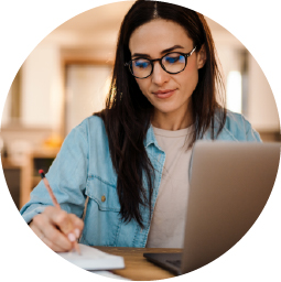 A woman with long dark hair and glasses writes in a notebook while using a laptop, seated at a desk in a well-lit room.