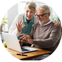 An older couple smiles and waves at a laptop screen while sitting at a table, suggesting they are video chatting with someone.
