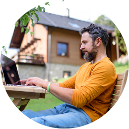 A man with a beard wearing an orange shirt sits at an outdoor wooden table, working on a laptop. A modern house and green lawn are visible in the background.